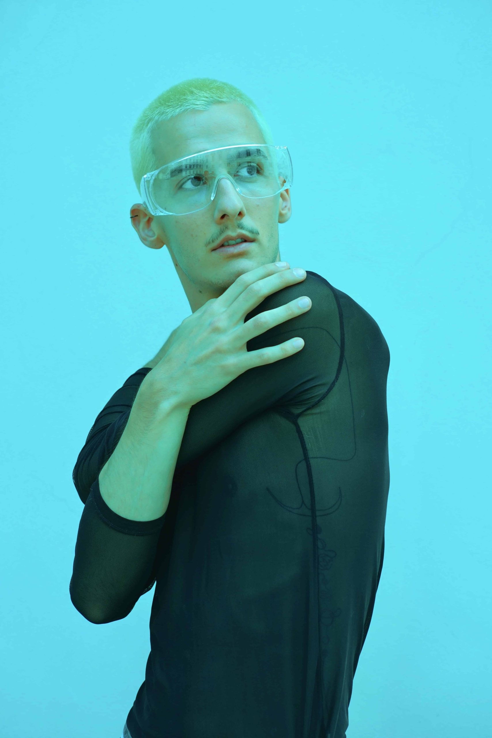 Ein blonder Mann in einem schwarzen Netz Oberteil steht vor einem grellblauen Hintergrund. Er trägt eine große transparente Brille und blickt zur Seite, seine Arme sind ineinander verschlungen.