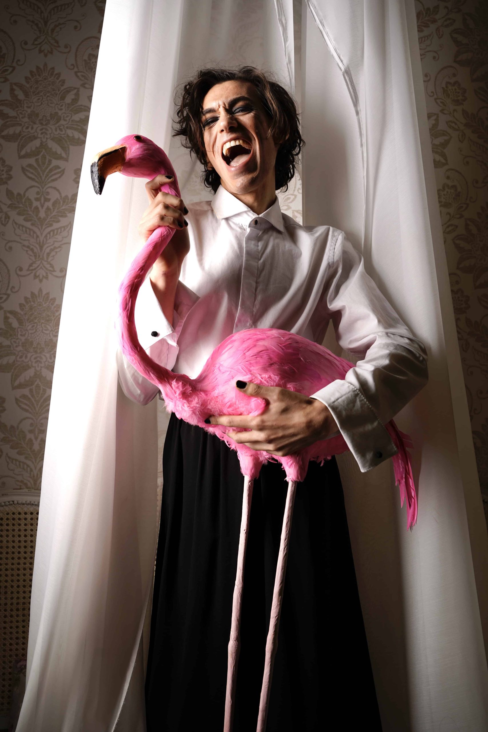 Ein mann steht vor einem weißen Vorhang und hält einen pinken Flamingo. er lacht mir weit geöffnetem Mund.